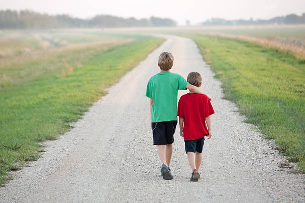 Two young boys walking arm in arm down a rural country gravel road. Prairie scene. Elementary aged caucasian children. Horizontal color image.